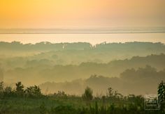 the sun is setting over some trees and fog in the distance, as seen from a hill