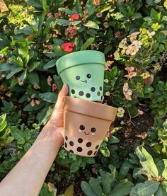 someone holding up two cups with faces painted on them in front of some bushes and flowers