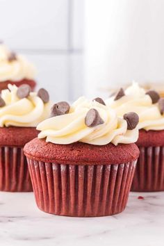 three red velvet cupcakes with white frosting and chocolate chips on top, sitting on a marble surface