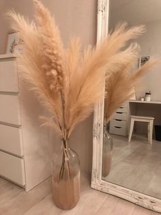 a vase filled with dry grass sitting next to a white dresser and mirror on top of a hard wood floor