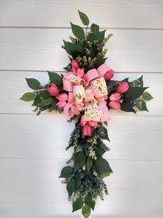 a cross decorated with pink flowers and greenery on a white wooden wall, in the shape of a cross