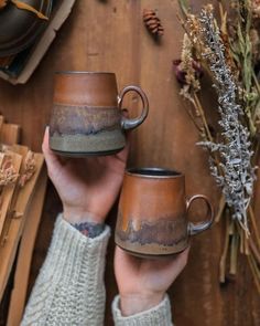 someone holding two coffee mugs in their hands on a wooden table with dried flowers