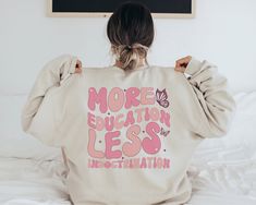 a woman sitting on top of a bed with her back to the camera, wearing a sweatshirt that says more education less information