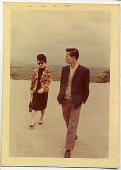 an old photo of two people walking in the sand with one man wearing sunglasses and another woman carrying a purse