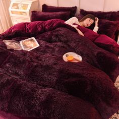 a woman laying in bed on top of a purple comforter with pillows and magazines