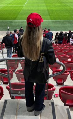 a woman with long hair wearing a red hat and black jacket standing in front of a stadium