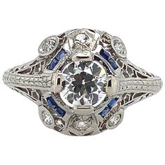 Art Deco platinum filigree ring featuring an European cut diamond weighing .62cts with GIA report. GIA report #2225056999 states H color and I1 clarity. The diamond measures 5.41mm x 5.47mm x 3.29mm. There are 8 specialty caliber French cut synthetic blue sapphire accents measuring about 1.5mm square, some have chips. There are 6 round brilliant cut diamond accents, 4 measure about 2.2mm and 2 measure about 2mm. The ring fits a size 8 3/4 finger and weighs 2.44dwt. It dates from the 1920's. Art Deco Gia Certified Sapphire Ring With Diamond, Art Deco Gia Certified Sapphire And Diamond Ring, Art Deco Sapphire Ring With Gia Certified Diamond, White Brilliant Cut Filigree Ring In Art Deco Style, White Filigree Ring In Brilliant Cut, Art Deco Style, Art Deco White Filigree Ring With Brilliant Cut, Gia Certified Diamond White Platinum Diamond Ring, Diamond White Sapphire Ring Platinum Brilliant Cut, Diamond White Platinum Sapphire Ring With Brilliant Cut