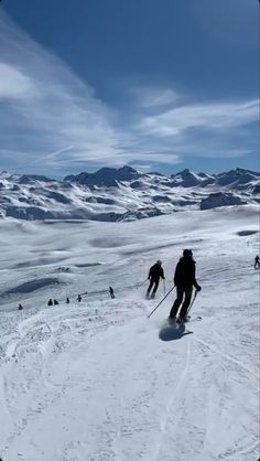 two people are skiing down a snowy mountain slope with mountains in the background and blue sky