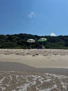 two umbrellas sitting on top of a sandy beach next to the ocean with trees in the background