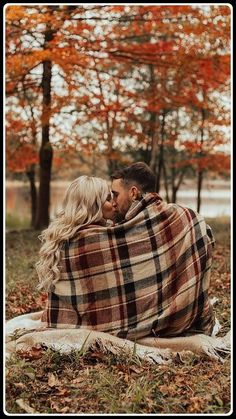 a man and woman wrapped up in a blanket sitting on the ground with trees behind them