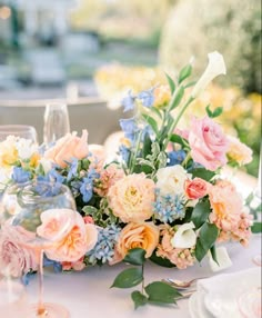 an arrangement of flowers and greenery on a table with wine glasses in the foreground