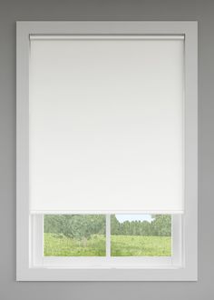 a window with white roller shades in front of a green field and trees behind it