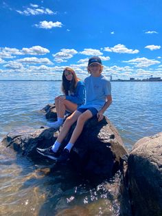 two young children sitting on rocks in the water near some rocks and blue sky with white clouds