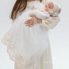 a woman holding a baby in her arms and wearing a white dress with flowers on it