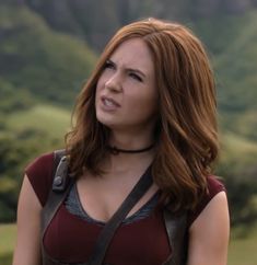 a woman with red hair wearing a brown top and black suspenders is standing in front of mountains