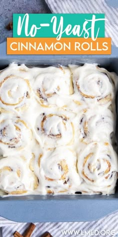 cinnamon rolls with icing in a blue baking pan on a striped towel next to cinnamon sticks