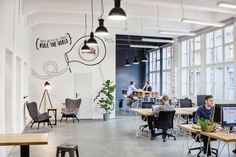 people working at desks in an office with large windows and hanging lights above them