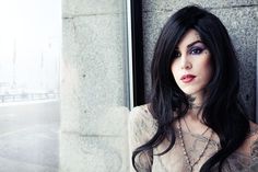 a woman with long black hair and piercings on her face leaning against a wall