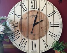 a wooden clock with roman numerals on the face next to a potted plant