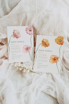 two wedding cards with watercolor flowers are laying on a white bed sheet and there is a flower in the middle