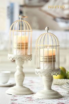 two white candles are sitting in bird cages on a table with fruit and teacups