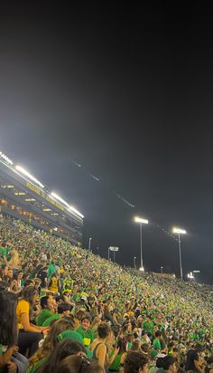 a large group of people sitting on top of a soccer field at night with lights in the background