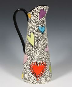 a ceramic pitcher with hearts painted on it's side and black handle, sitting against a gray background