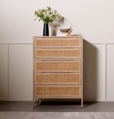 a wicker chest of drawers with flowers in a black vase on top and a white wall behind it