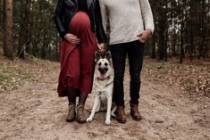 two people and a dog standing on a dirt path in the woods with trees behind them