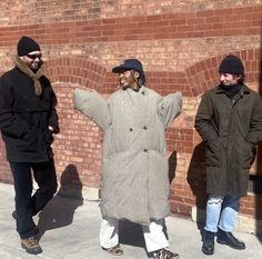 three men standing next to each other in front of a brick wall and wearing coats