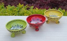 three colorful bowls sitting on top of a white table cloth next to green plants and bushes