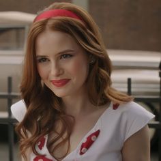 a woman with long red hair wearing a white shirt and polka dot headband smiling at the camera