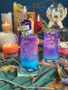 two glasses filled with blue and purple liquid sitting on top of a table next to candles