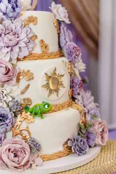 a three tiered wedding cake with purple flowers on the side and a green frog sitting on top