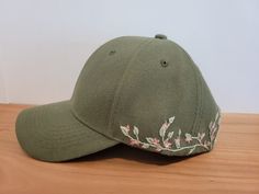 a green baseball cap with pink flowers embroidered on the front and side, sitting on a wooden surface