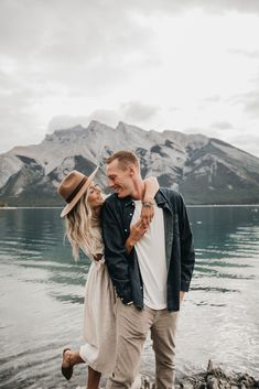 a man and woman standing next to each other near the water with mountains in the background