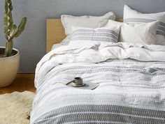 a bed with white sheets and pillows next to a cacti in a pot