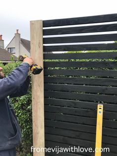 a man working on a wooden fence in front of some bushes and trees with a measuring tape