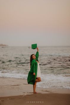 a woman in a green graduation cap and gown on the beach with her arms up