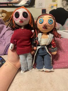 two dolls sitting on top of a bed next to each other in front of a person's hand