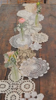 several vases with flowers are lined up on an old wooden table, along with doily