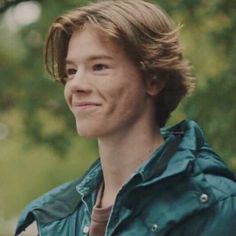 a woman in a green jacket smiling at the camera with trees in the back ground