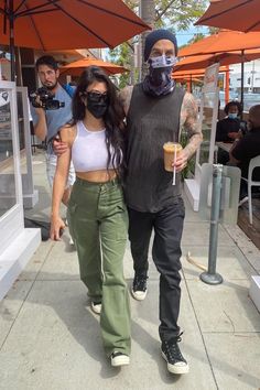 a man and woman walking down the sidewalk with masks on their faces while holding drinks