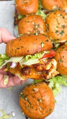 a hand is holding a sandwich with lettuce and tomato on it, while other sliders are in the background