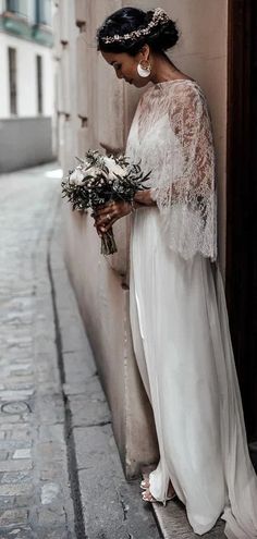 a woman standing next to a wall holding flowers and wearing a white dress with sheer sleeves