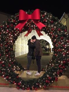 two people standing in front of a christmas wreath with red bows and lights on it