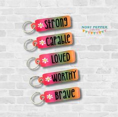 three key chains with flowers on them hanging from a brick wall, one saying strong carable loved worthy brave