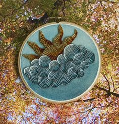 an embroidered sun and clouds on a blue hoop hanging from a tree branch with autumn leaves in the background