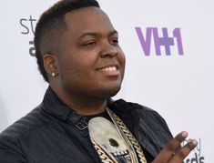 a man in black jacket standing next to a white wall with his hands out and smiling at the camera