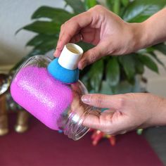 two hands holding a pink and blue bottle with something in it that is on top of a table